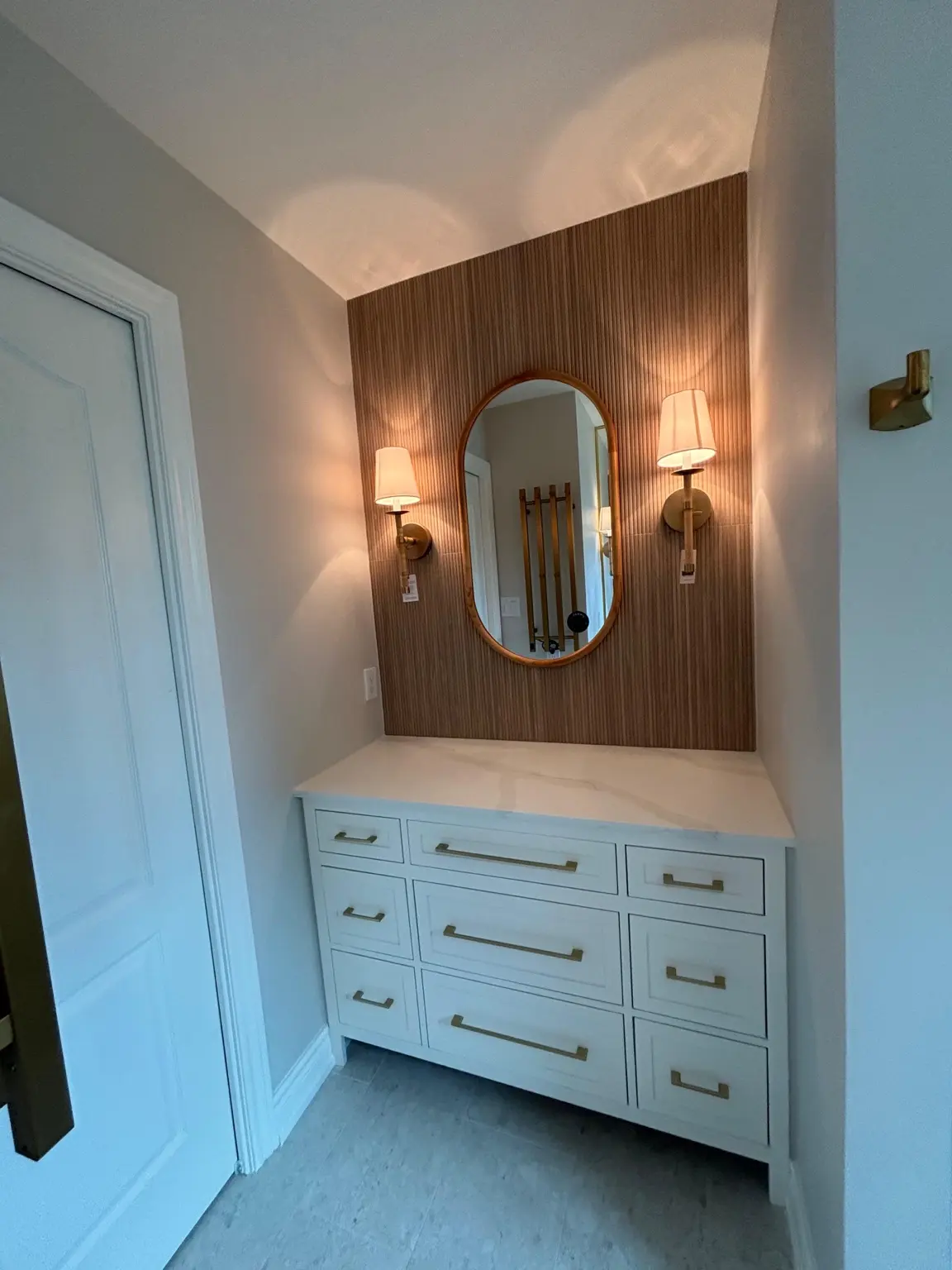 Custom white bathroom vanity with warm wood accent wall, oval mirror and brass sconces